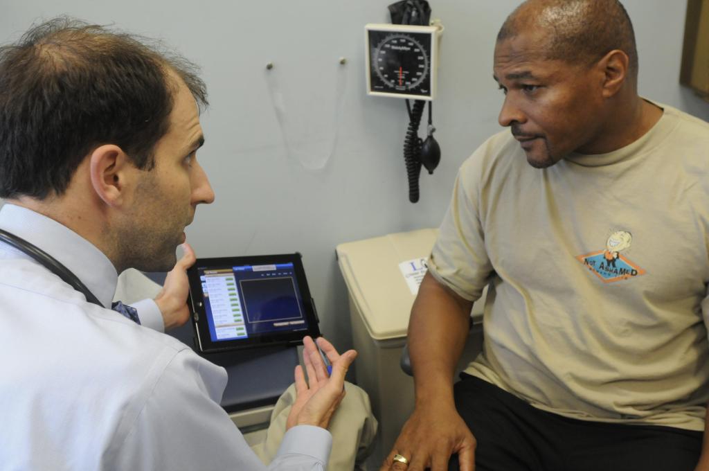 Clinician and patient sitting across from each other in a VA exam room with an Ambient Scribe screen visible.